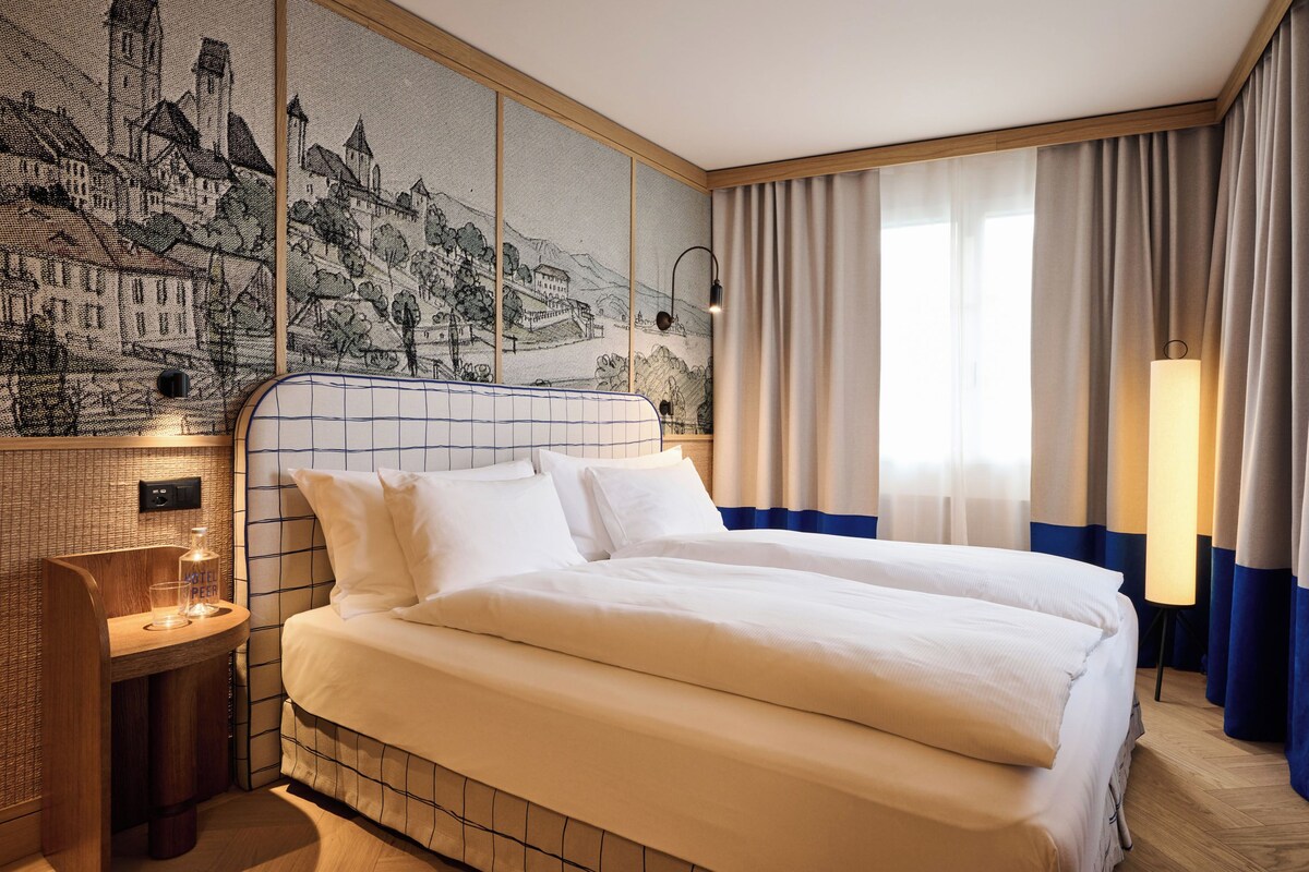 Una habitación de hotel con una cama grande, una mesita de noche pequeña con una botella y un vaso, una lámpara de pared y un ventanal con cortinas.