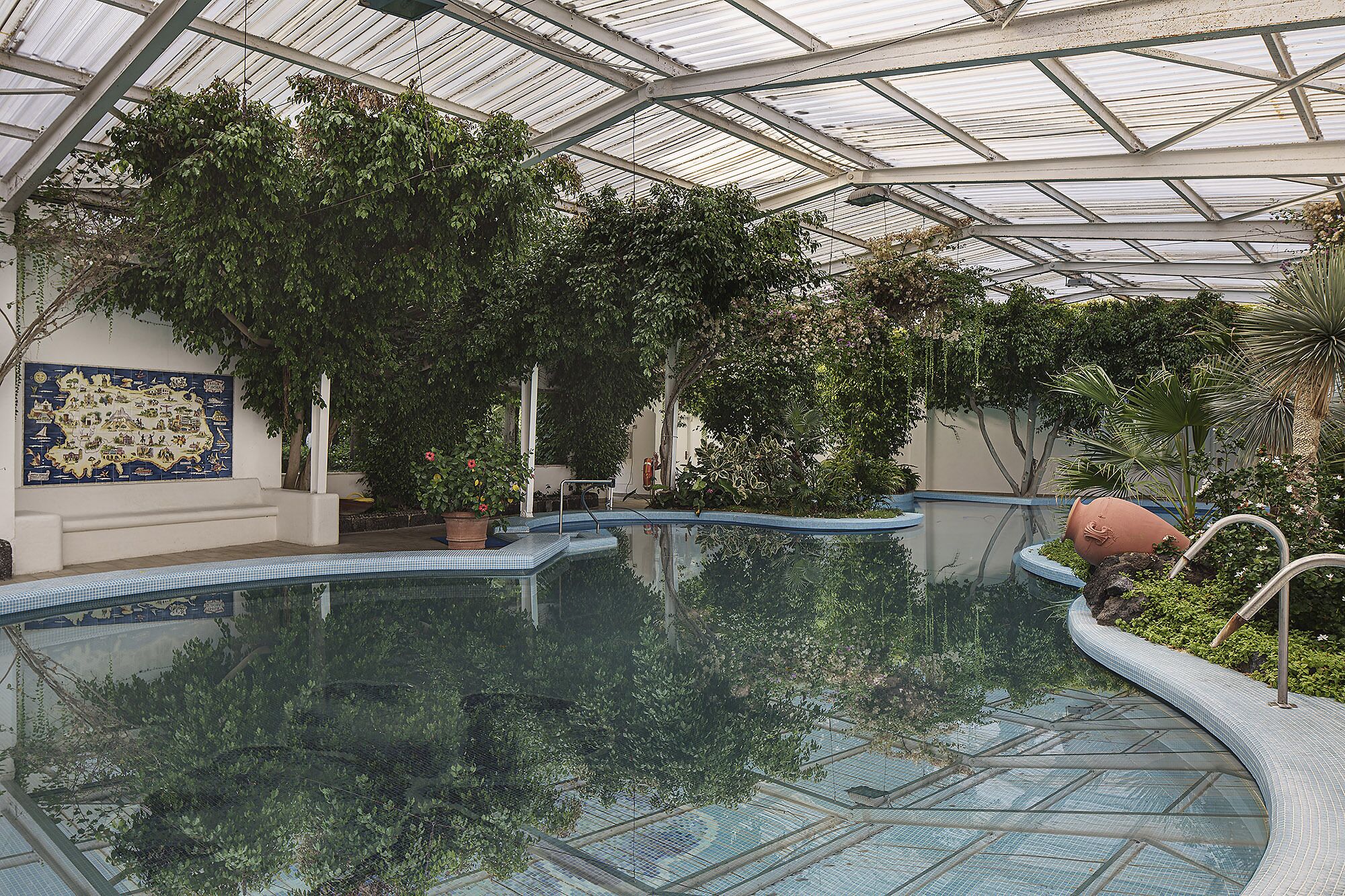 Una piscina interior con techo de cristal, rodeada de abundante vegetación y una pared decorativa de azulejos.