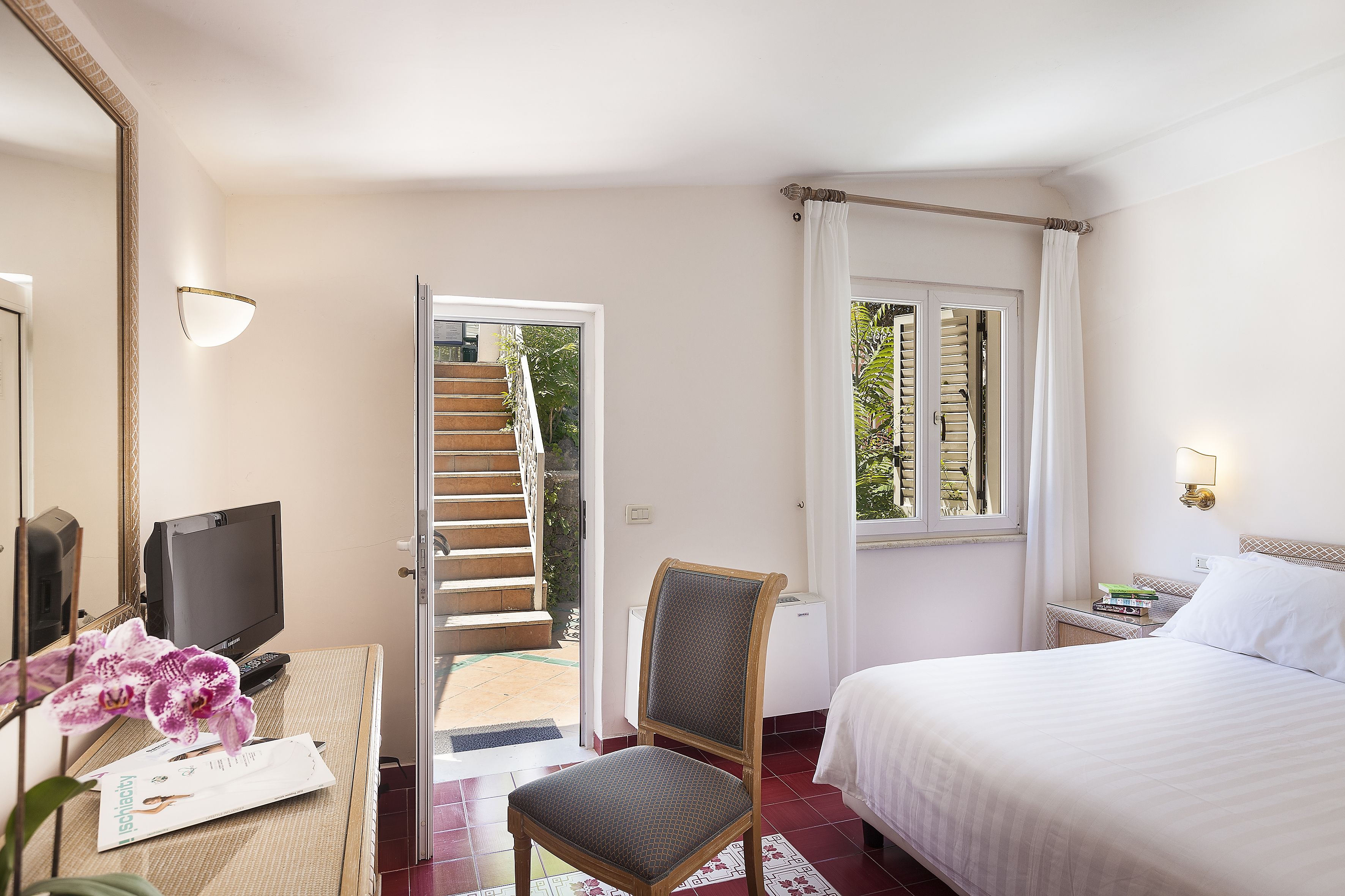 A hotel room with a bed, a desk with a computer, a chair, a window with curtains, and a view of a staircase leading to an outdoor area.