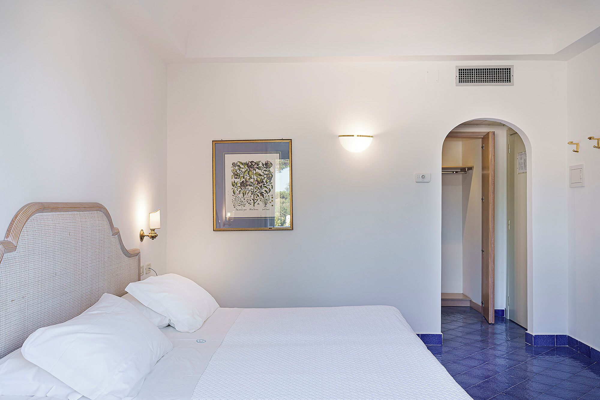 Habitación de hotel con cama, una ilustración botánica enmarcada, una lámpara de pared y una puerta que da al baño.