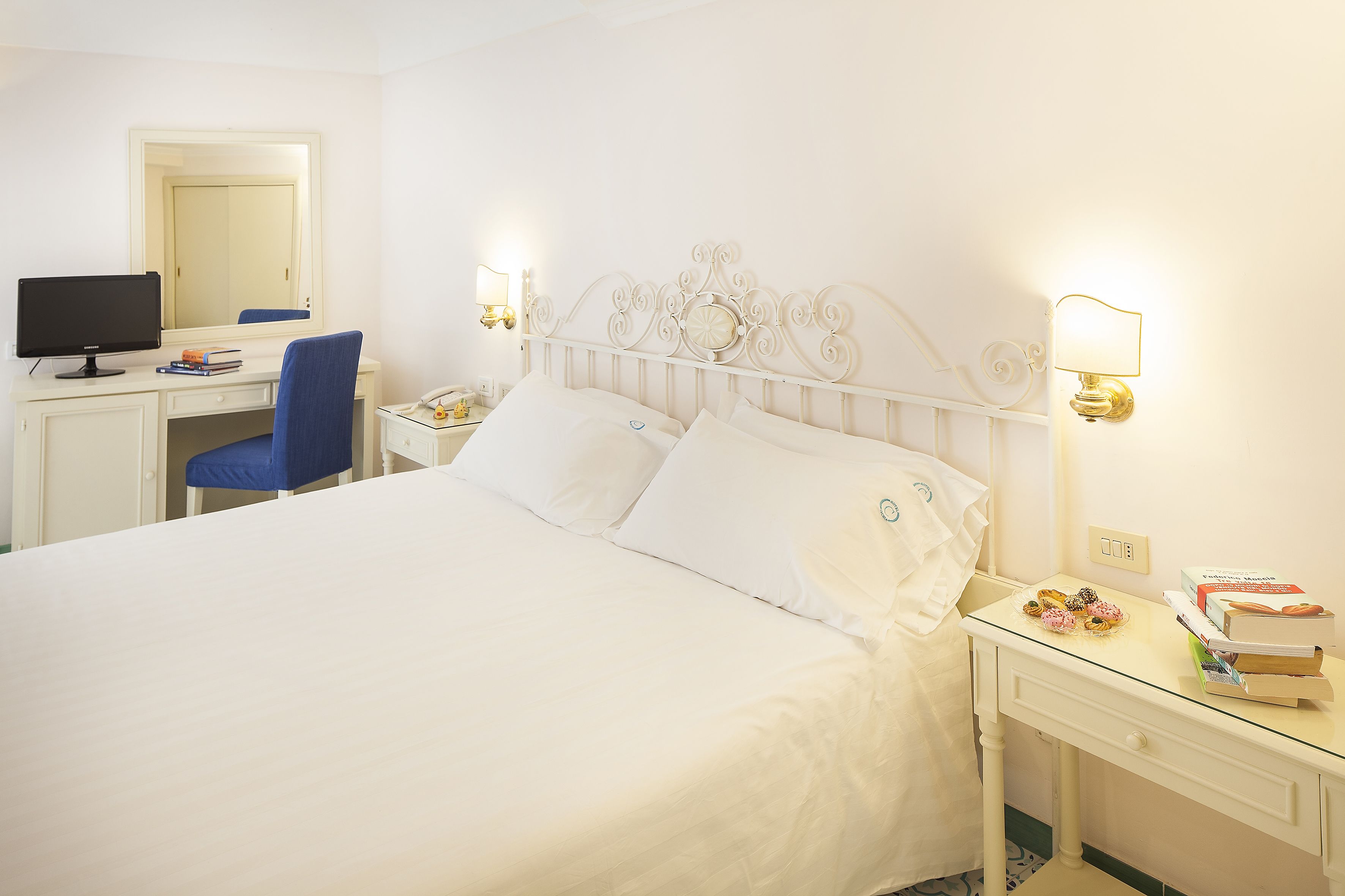 Una camera d'albergo con un letto, una scrivania con un computer, una sedia e comodini con lampade e libri.