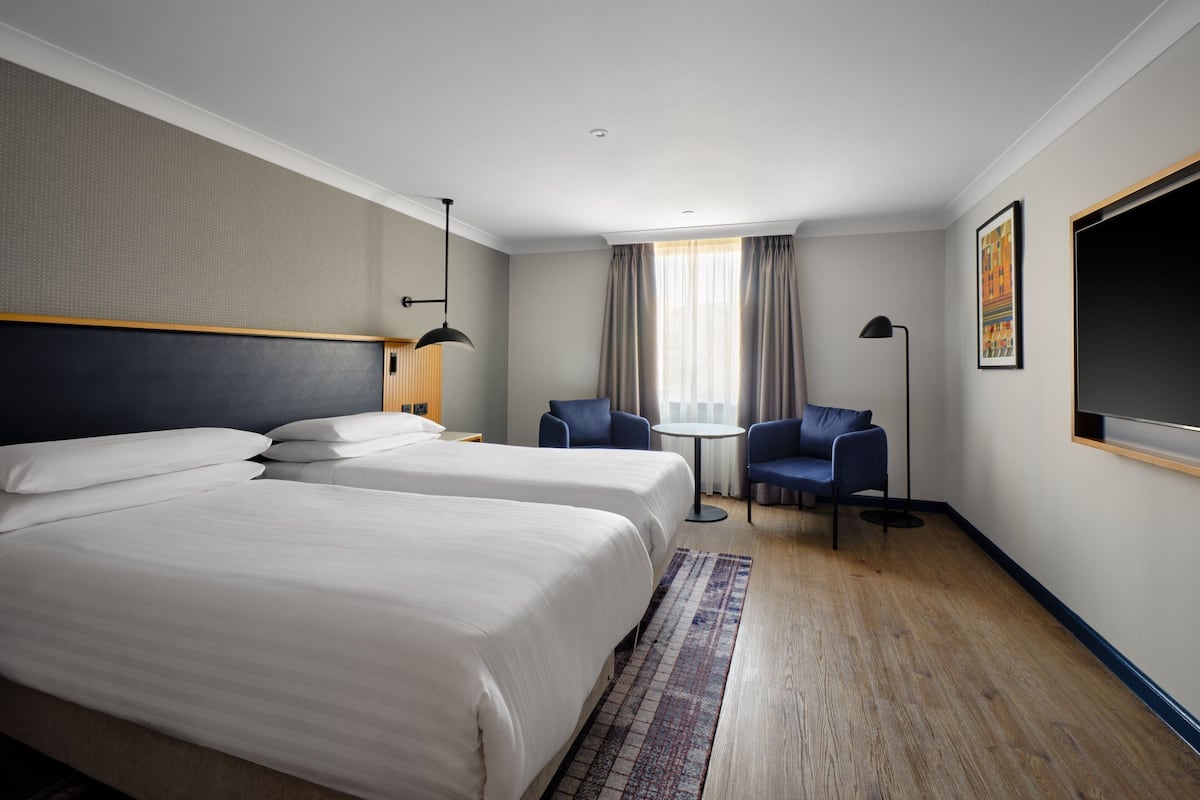 Habitación de hotel con dos camas, una mesita pequeña, dos sillas, televisor y ventana con cortinas.
