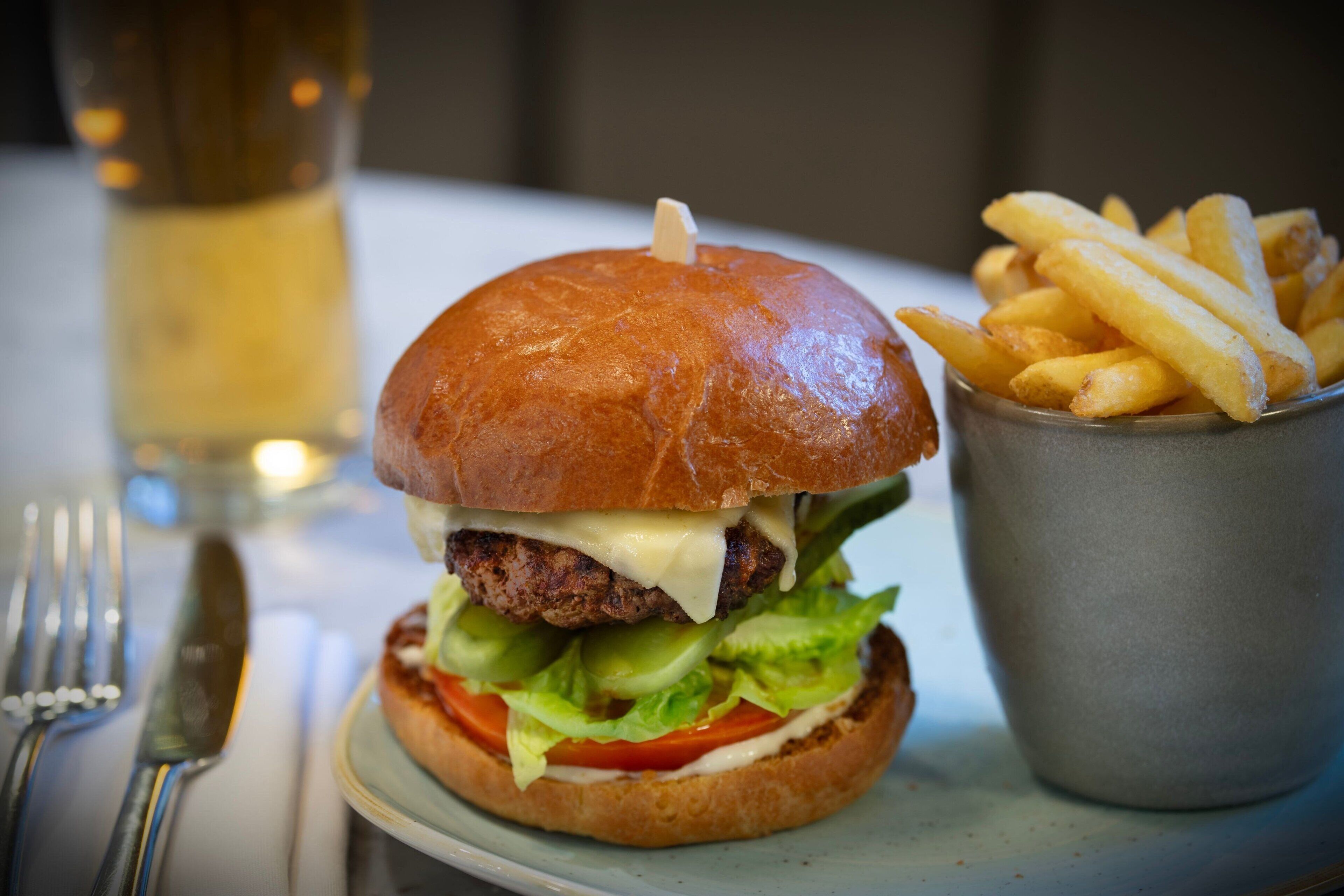 A burger with fries on a plate, a glass of beer, and cutlery on a table.