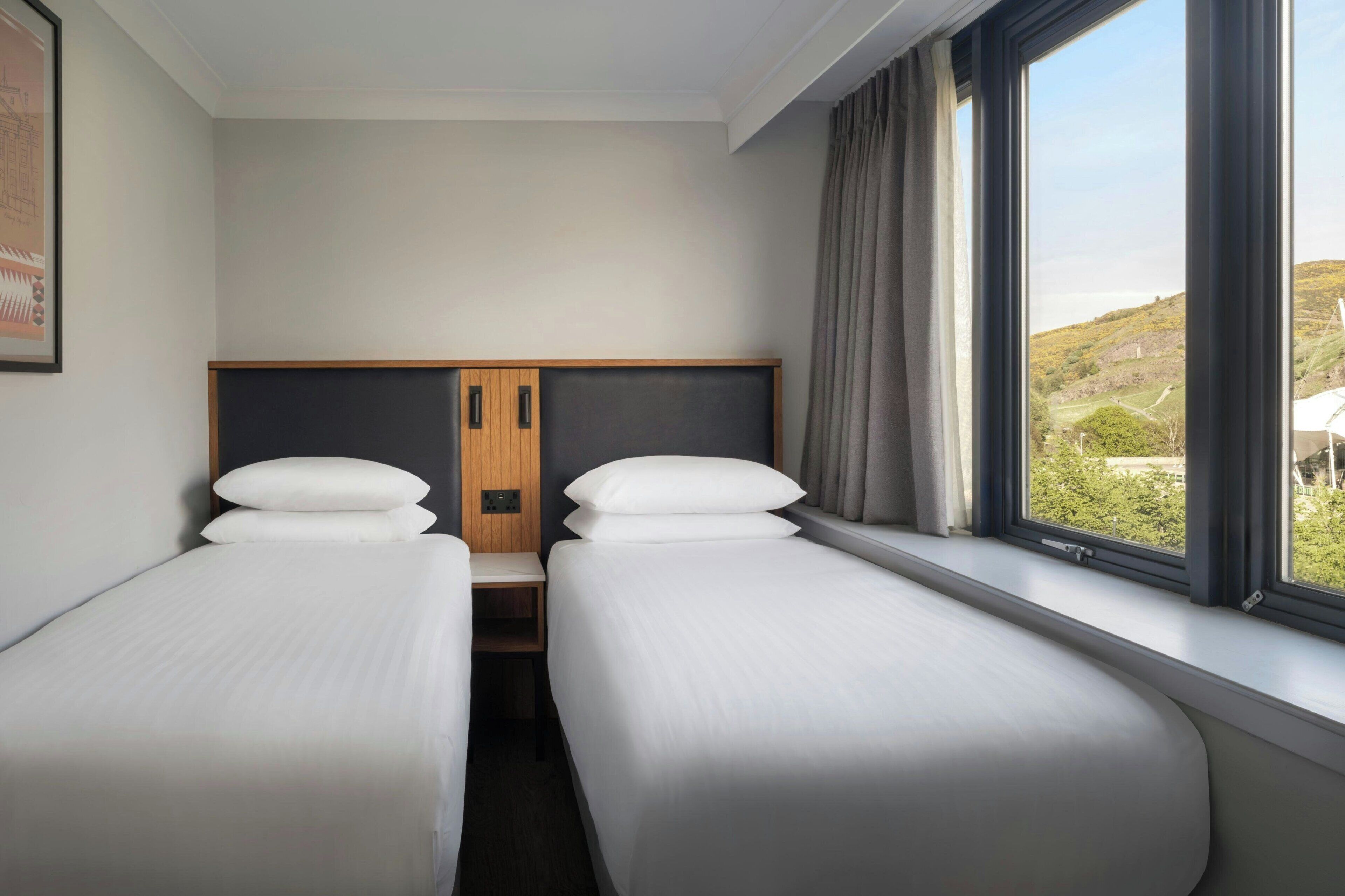 A hotel room with two single beds, a wooden headboard, a window with a view of greenery, and a framed picture on the wall.