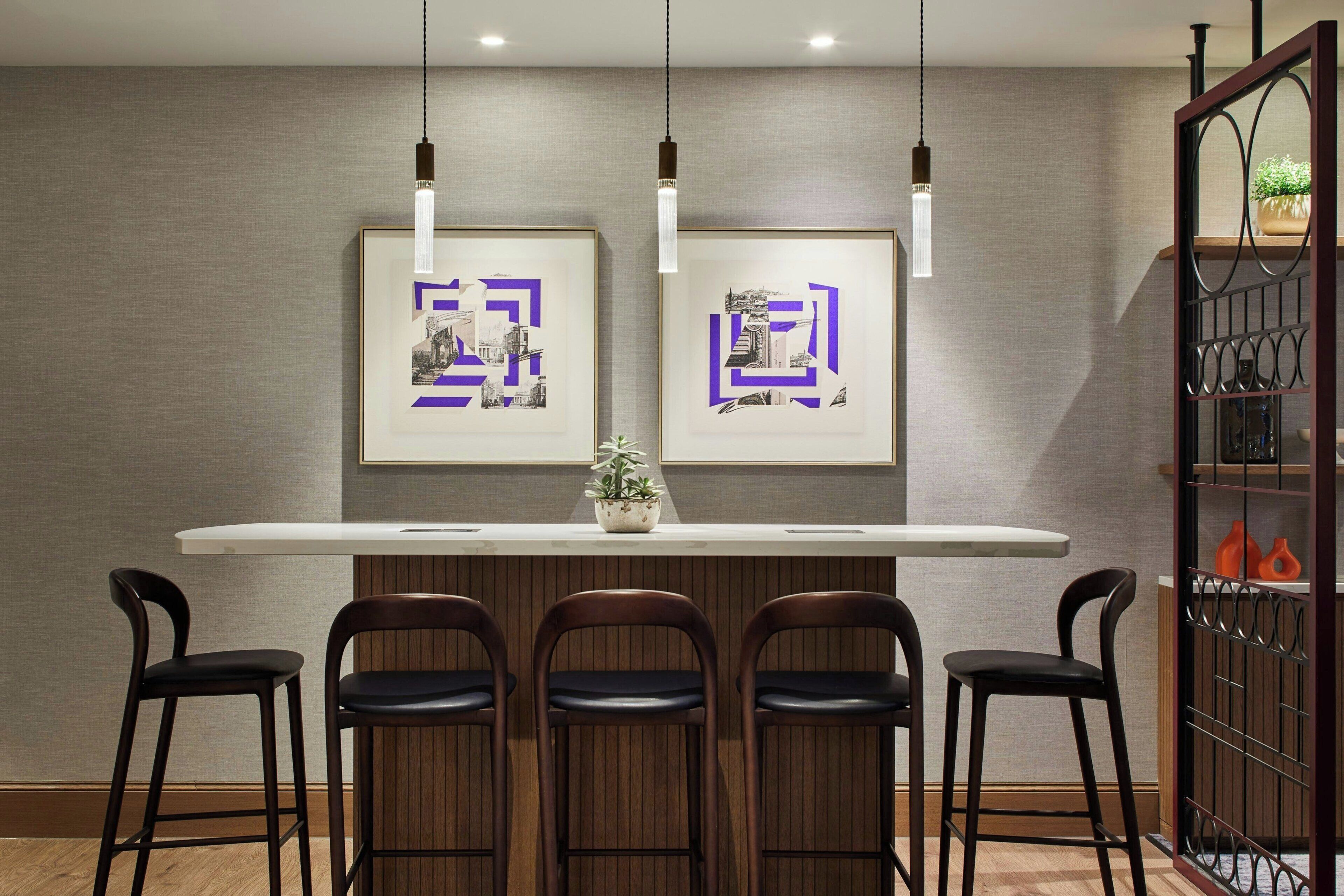 A modern kitchen with a wooden bar counter, three bar stools, pendant lights, and framed artwork on the wall.