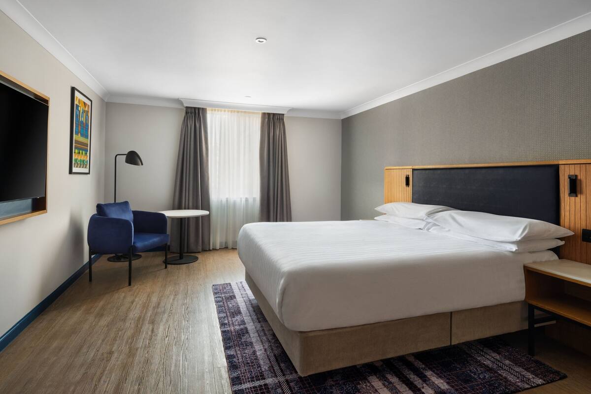Una habitación de hotel moderna con una cama grande, un escritorio, una silla, un televisor y una ventana con cortinas.