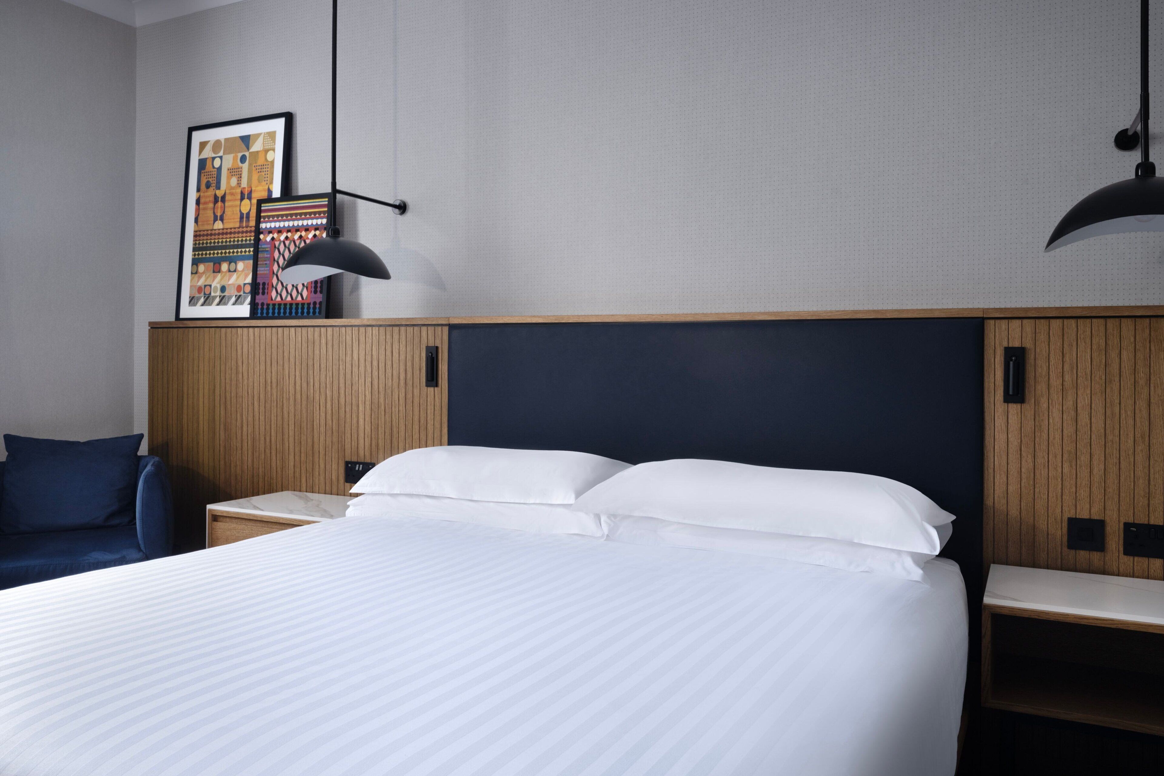 A hotel room with a large bed, wooden headboard, two bedside tables, a nightstand, a framed picture on the wall, and a pendant light.