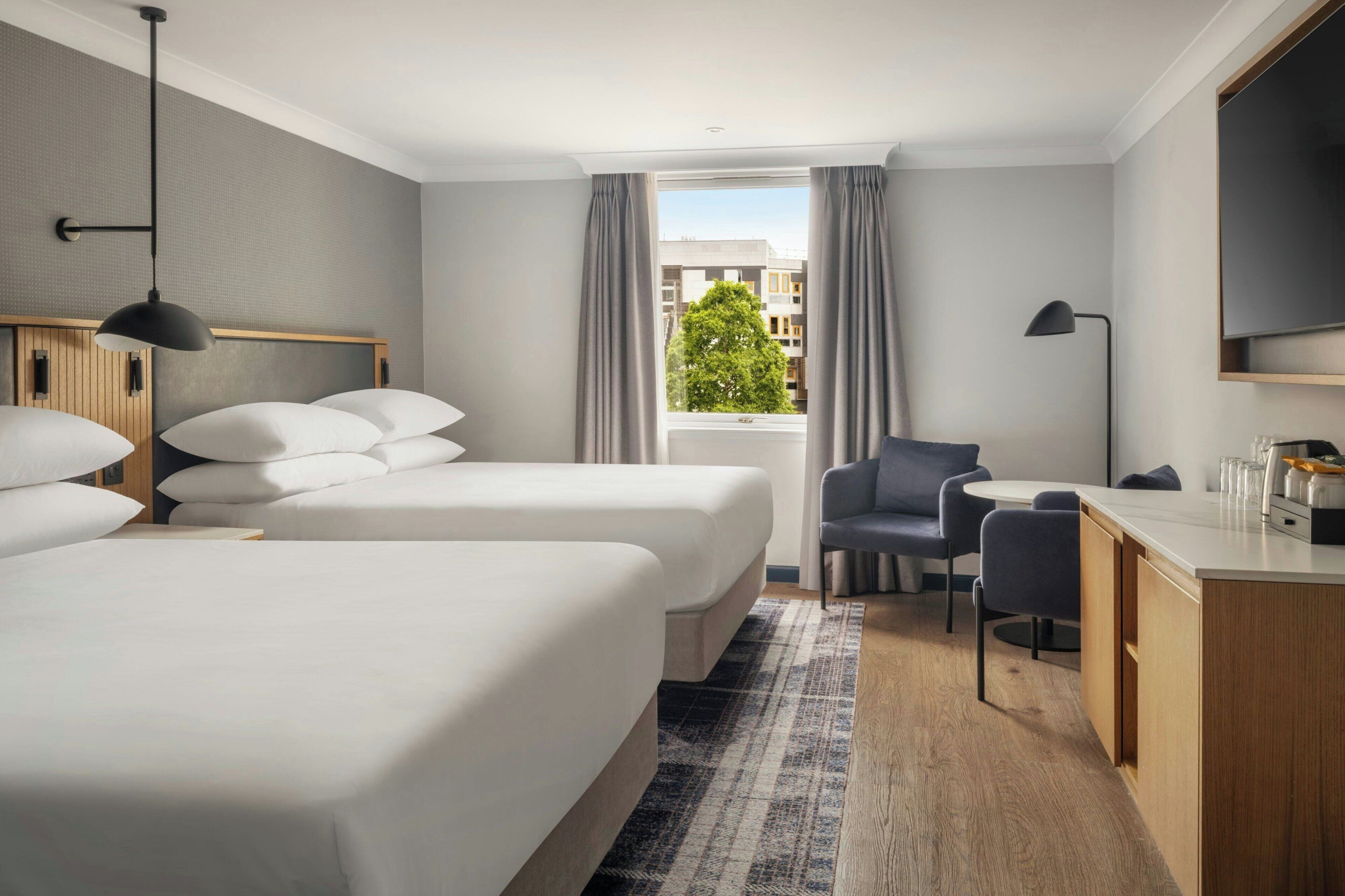 A hotel room with two beds, a desk, a chair, a window with curtains, and a view of buildings and a tree.