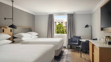 A hotel room with two beds, a desk, a chair, a window with curtains, and a view of buildings and a tree.