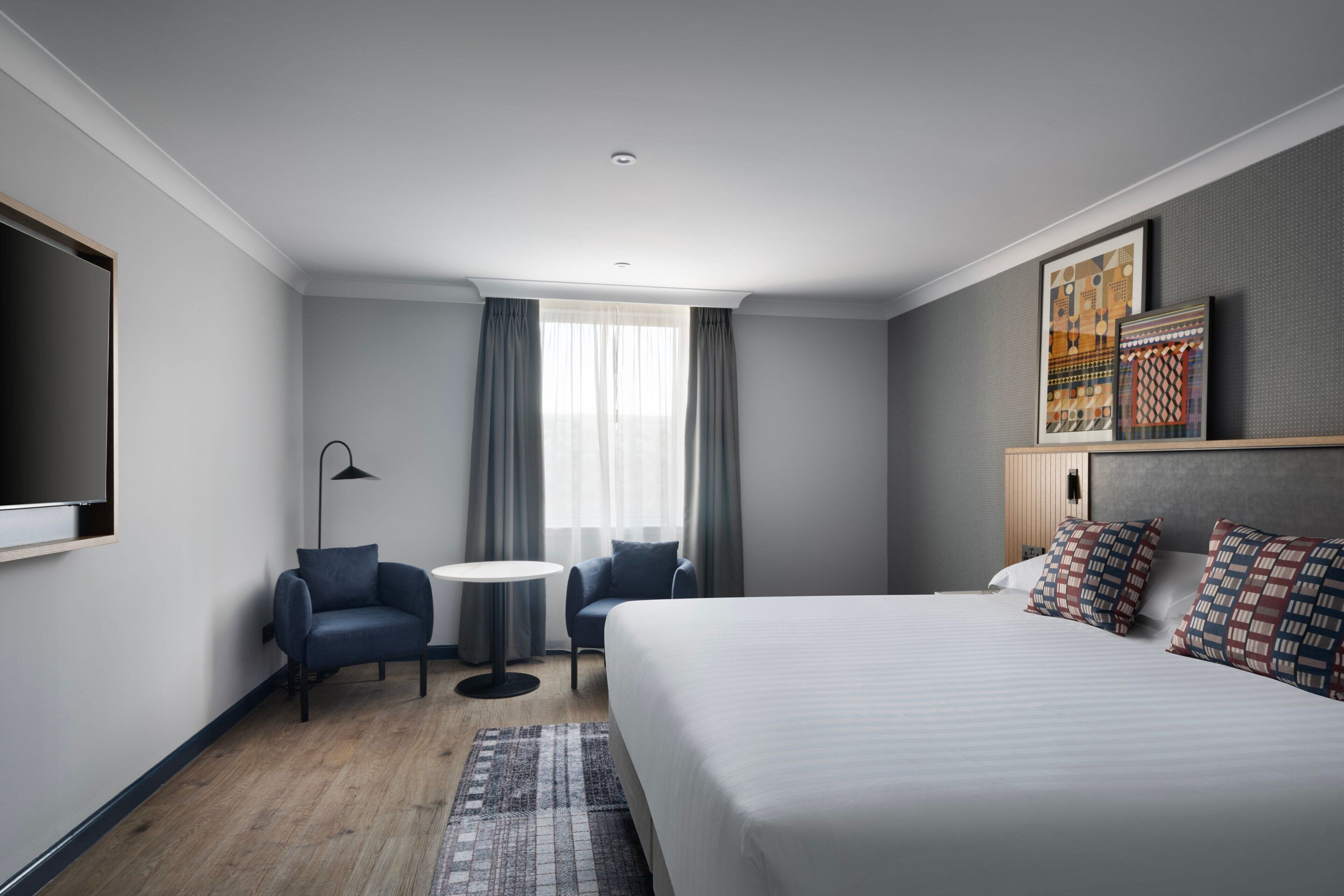 A modern hotel room with a large bed, a desk, two chairs, a small round table, and a TV mounted on the wall.