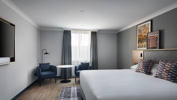 A modern hotel room with a large bed, a desk, two chairs, a small round table, and a TV mounted on the wall.