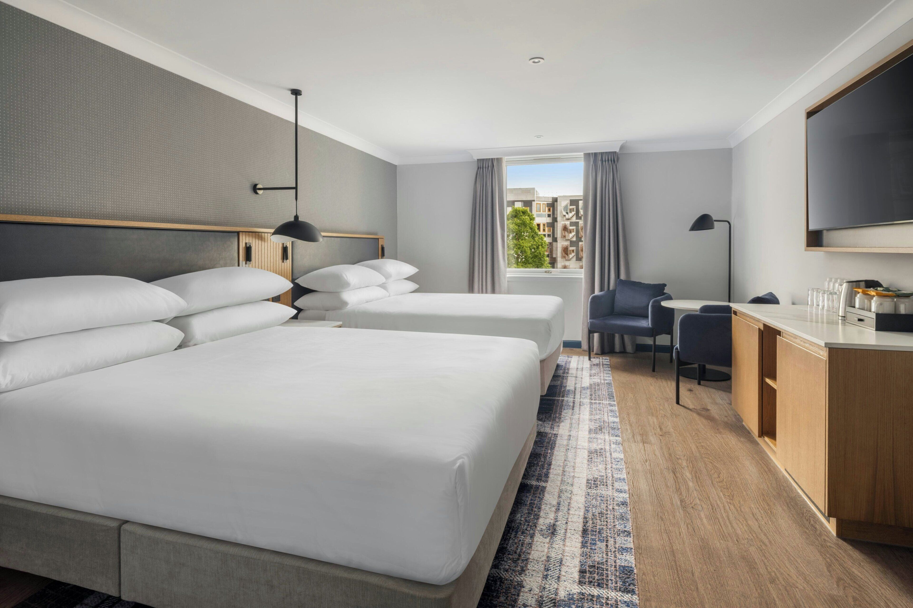 A hotel room with two beds, a desk, a chair, a TV, and a window with a view of buildings.