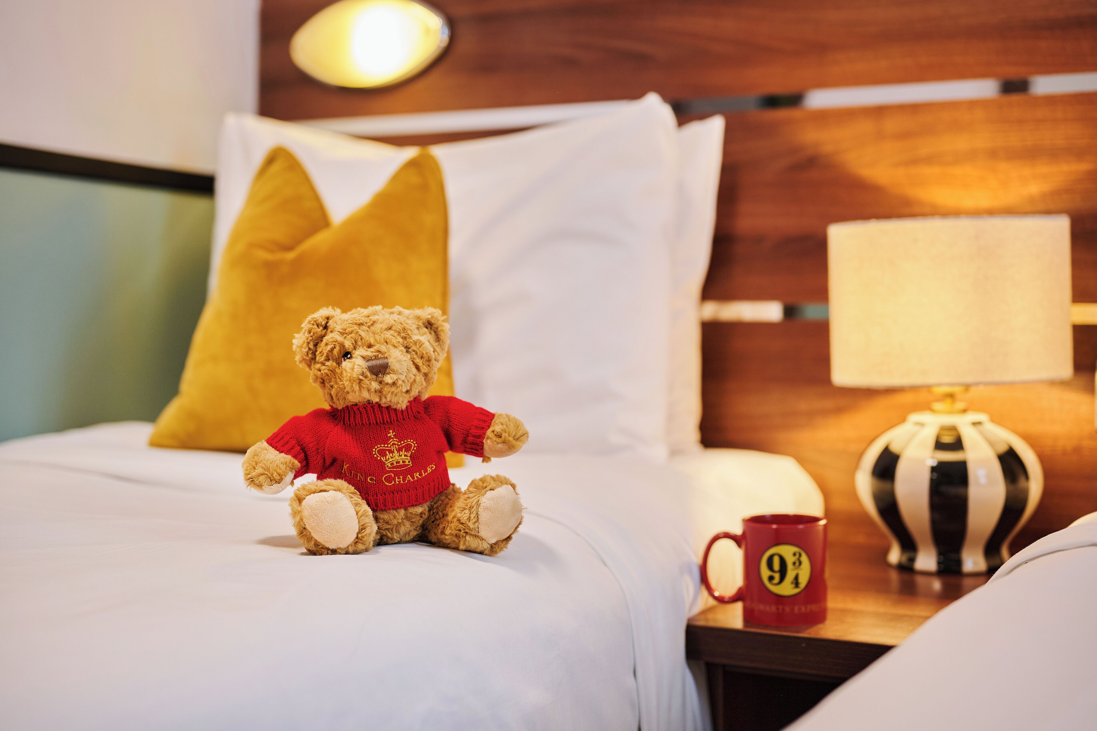 A teddy bear on a bed with a yellow pillow, a red mug with a number, and a lamp on a bedside table.