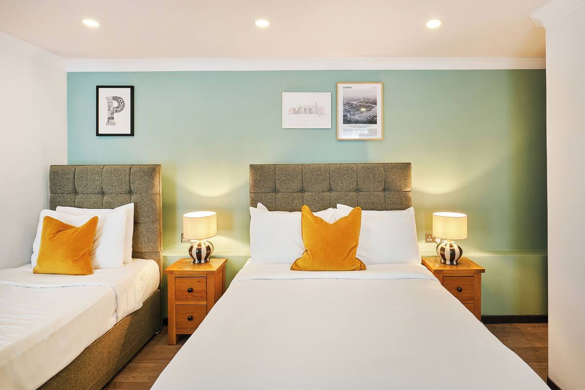 Une chambre d’hôtel avec deux lits, chacun doté d’une tête de lit, de draps blancs et d’un seul oreiller jaune. Il y a deux tables de chevet avec des lampes, et les murs sont peints dans une couleur vert d’eau pâle.