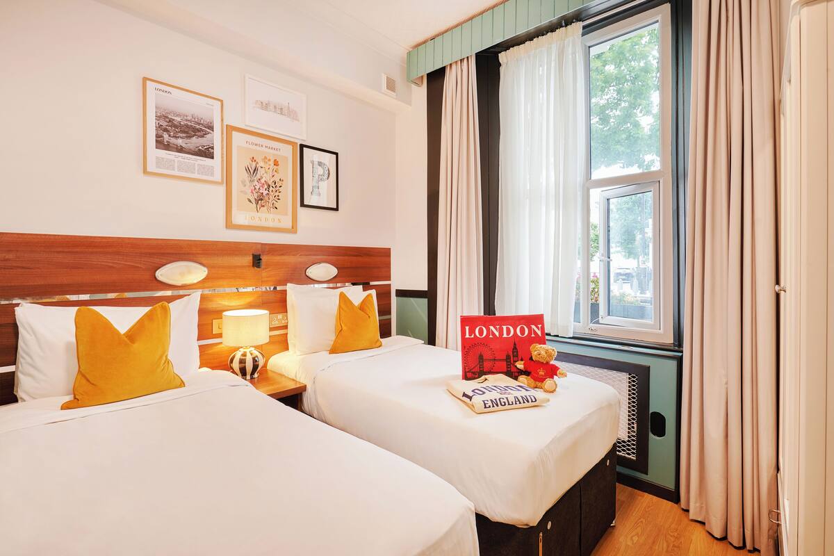 Une chambre d’hôtel avec deux lits, une table de chevet avec une lampe, des tableaux encadrés au mur et une fenêtre avec des rideaux.