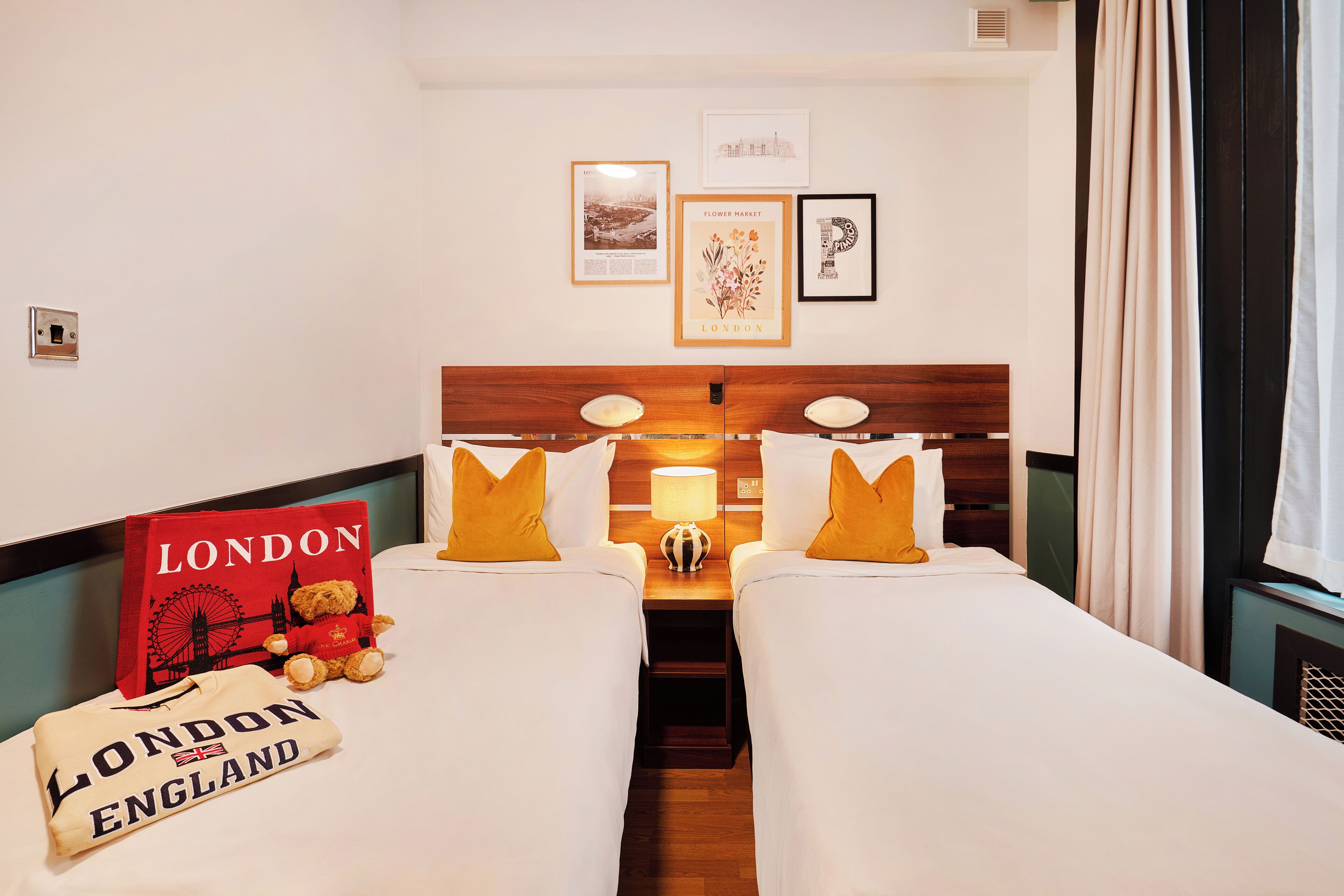 A hotel room with two beds, a nightstand with a lamp, a teddy bear, and a 'London' themed pillow.