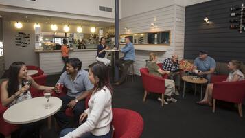 A social gathering in a modern bar area with round tables, red chairs, and a bar counter with various bottles.