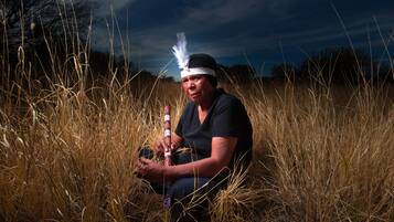 A person sitting in tall grass holding a traditional flute.