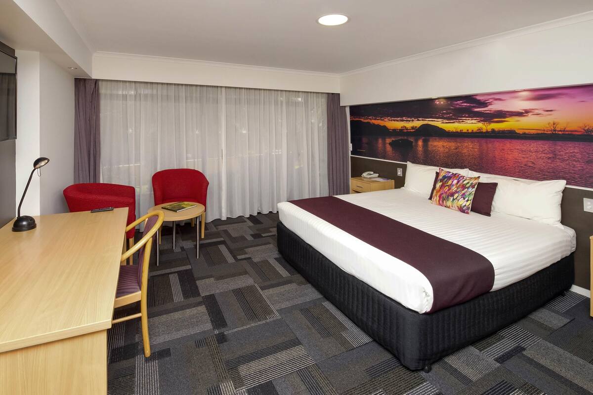 A hotel room with a large bed, a desk, a chair, a small table, a nightstand, and a painting of a sunset over water.