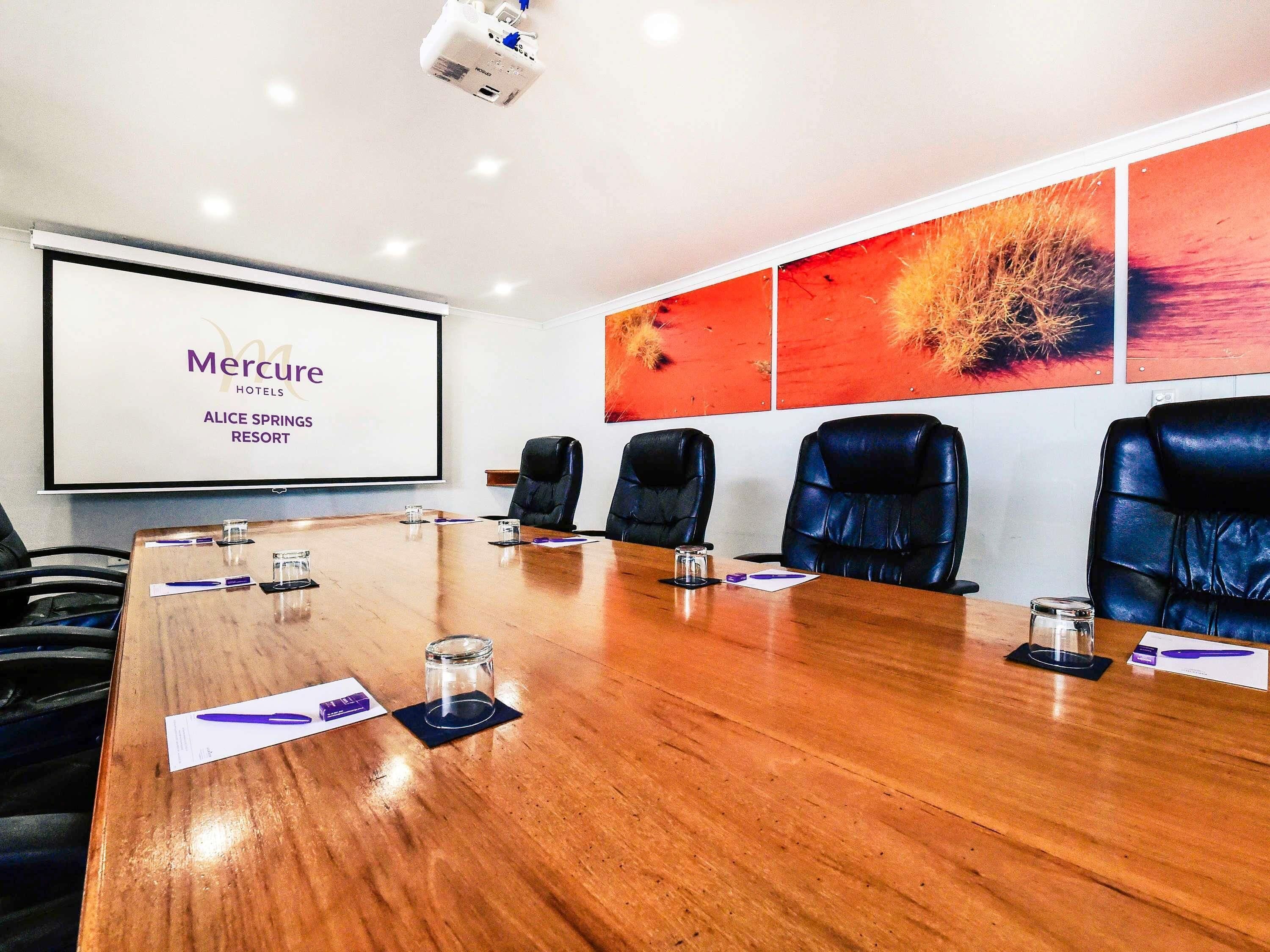 A conference room with a long table, black leather chairs, a projector screen, and a large painting on the wall.