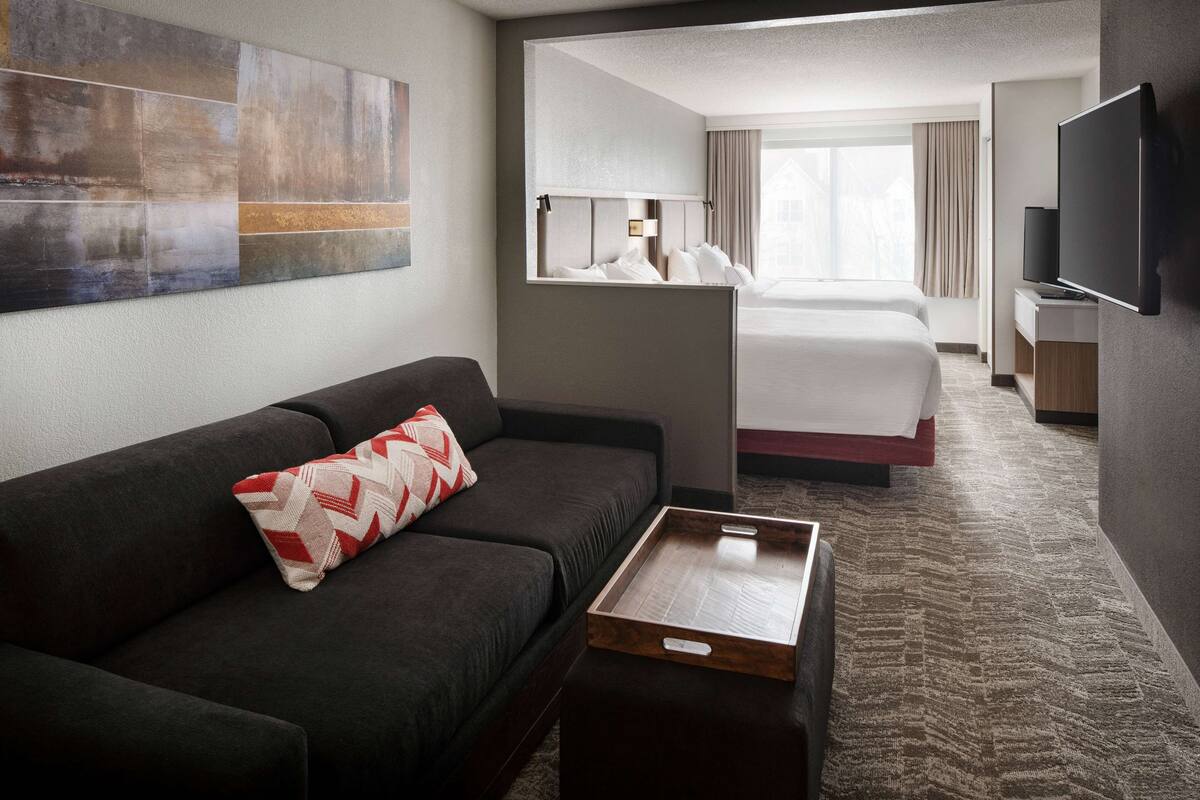A hotel room with a dark sofa, a wooden coffee table, a bed with white linens, a TV, and a large abstract painting on the wall.
