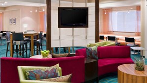 A hotel lobby with red and green sofas, a flat-screen TV, a dining area, and a large window with curtains.