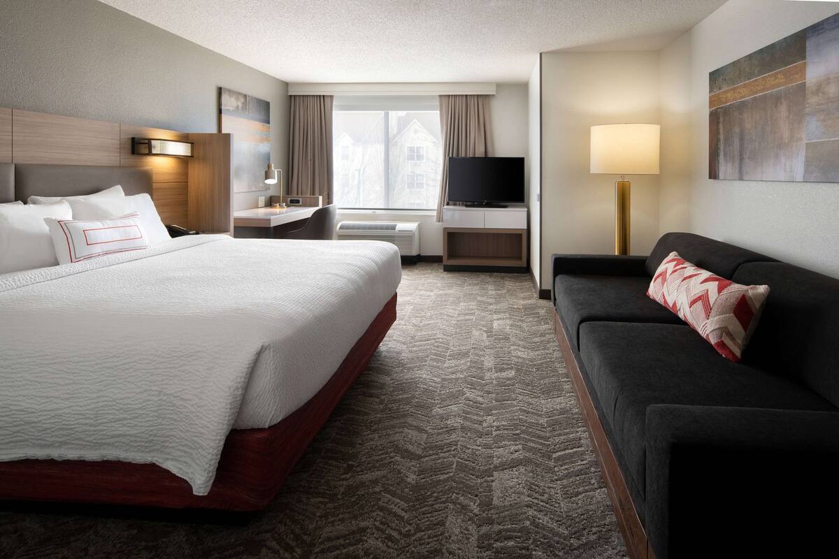 A hotel room with a large bed, a sofa, a desk with a lamp, a TV, and a window with curtains.