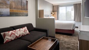A modern hotel room with a dark sofa, a wooden tray table, a bed with a white mattress, a large window with curtains, and a wall-mounted TV.