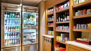 A refrigerated display with various beverages and snacks, wooden shelves stocked with packaged goods, and a counter area.