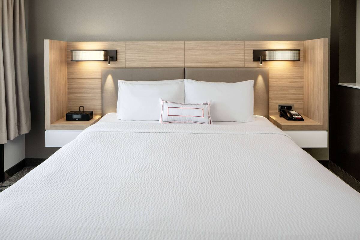 A hotel room with a large bed, two bedside tables, a nightstand, a lamp, and a wall-mounted light fixture.
