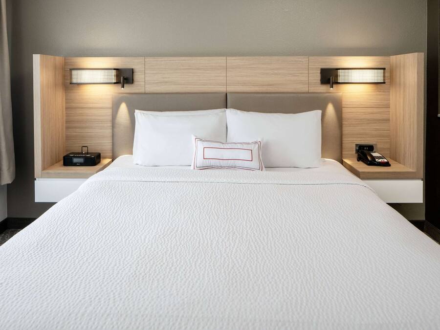 A hotel room with a large bed, two bedside tables, a nightstand, a lamp, and a wall-mounted light fixture.