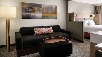 Ein Hotelzimmer mit einem dunkelgrauen Sofa, einem Holznachttisch, einer Stehlampe, einem Bett mit weißem Kopfteil und einem großen abstrakten Gemälde an der Wand.