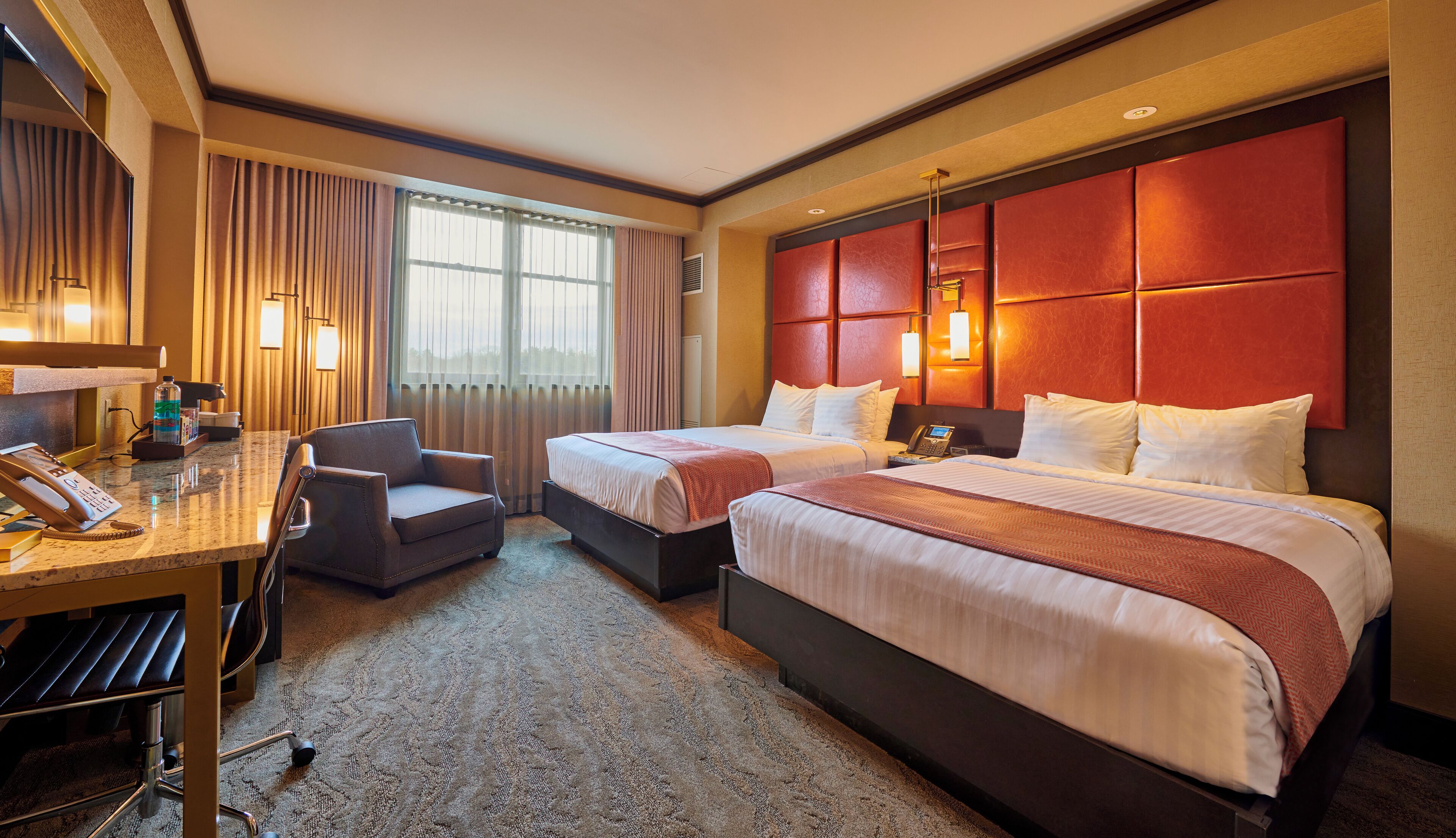 A hotel room with two beds, a desk with a telephone, a chair, and a large window with curtains.