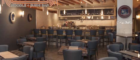 Interior de un restaurante con mesas y sillas, una zona de cocina y un letrero que dice 'LUCKY8 Noodle + Sushi Bar'.