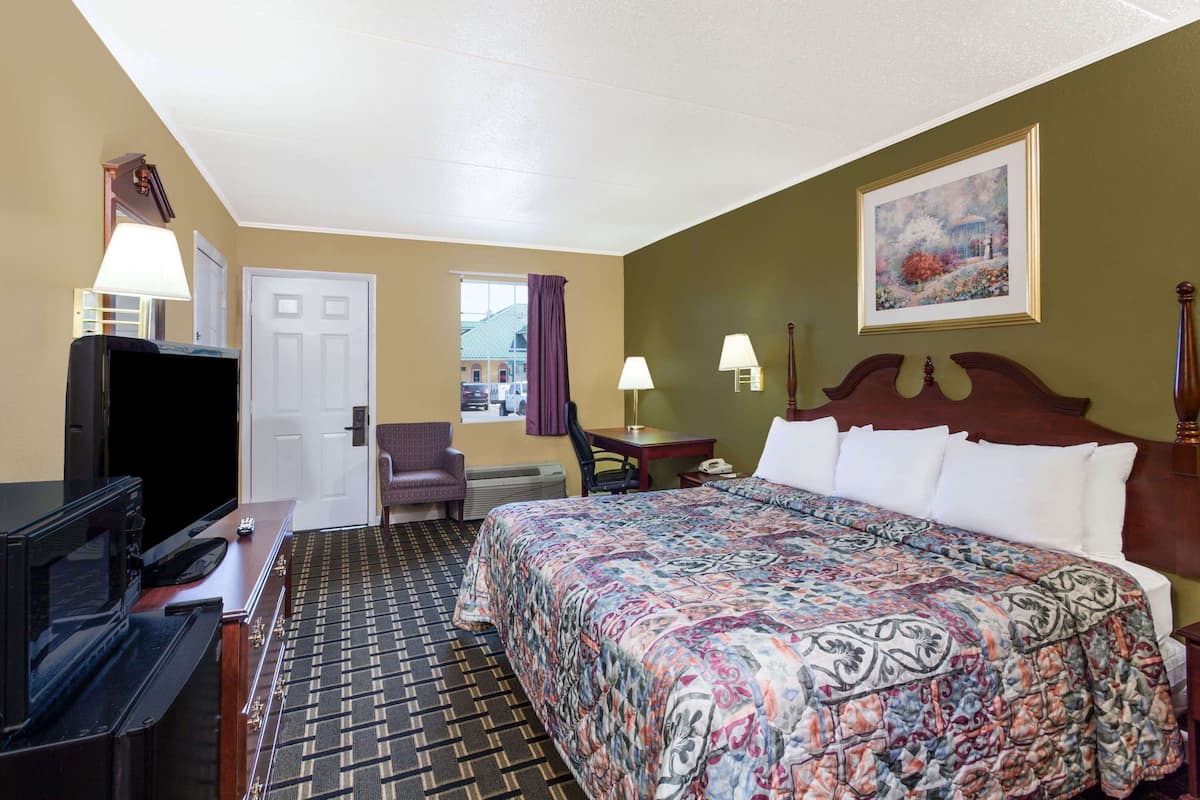 Habitación de hotel con cama, escritorio con televisor, silla, ventana con cortinas y un cuadro en la pared.