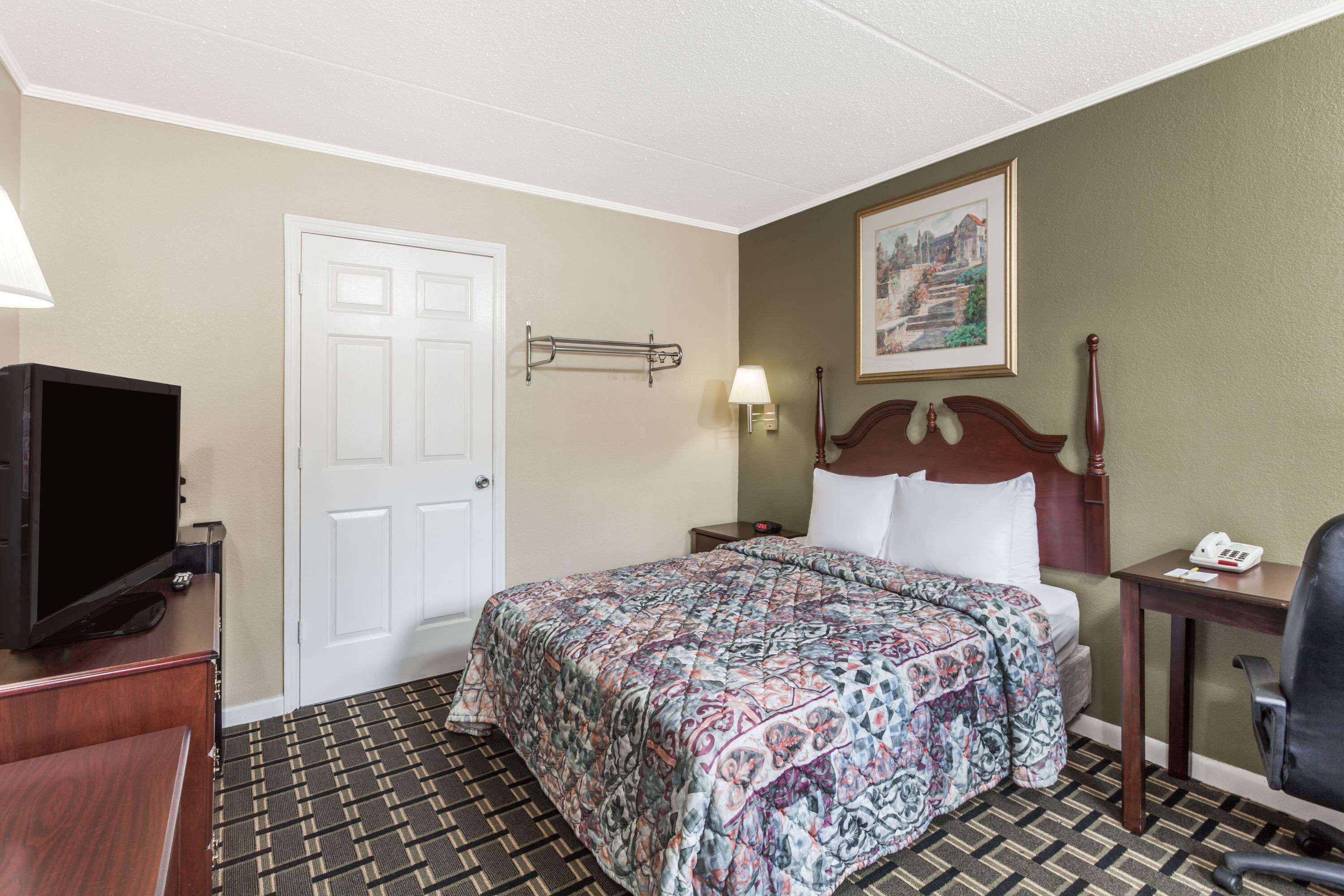 A hotel room with a bed, a television, a desk with a lamp, a chair, and a painting on the wall.