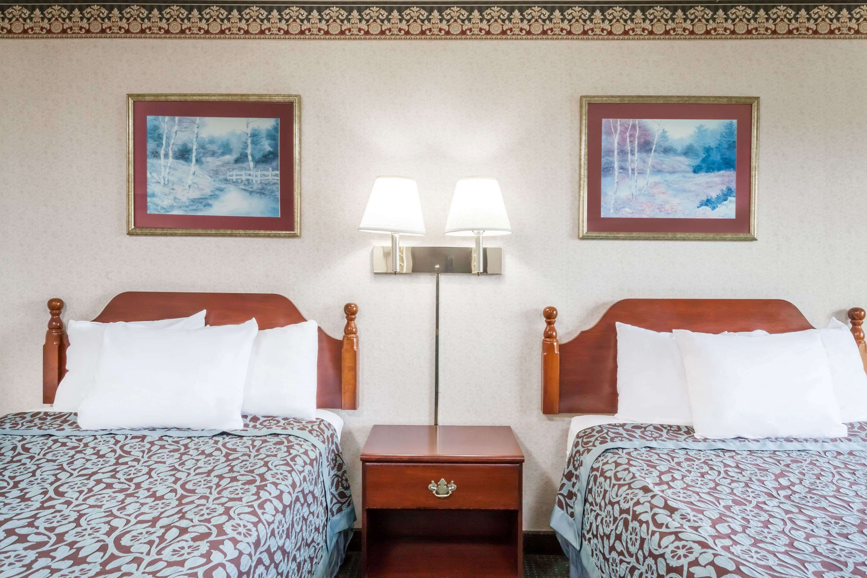 Habitación de hotel con dos camas, cada una con cabecero de madera y ropa de cama estampada. Entre las camas hay dos lámparas fijadas a la pared, y dos cuadros enmarcados encima.