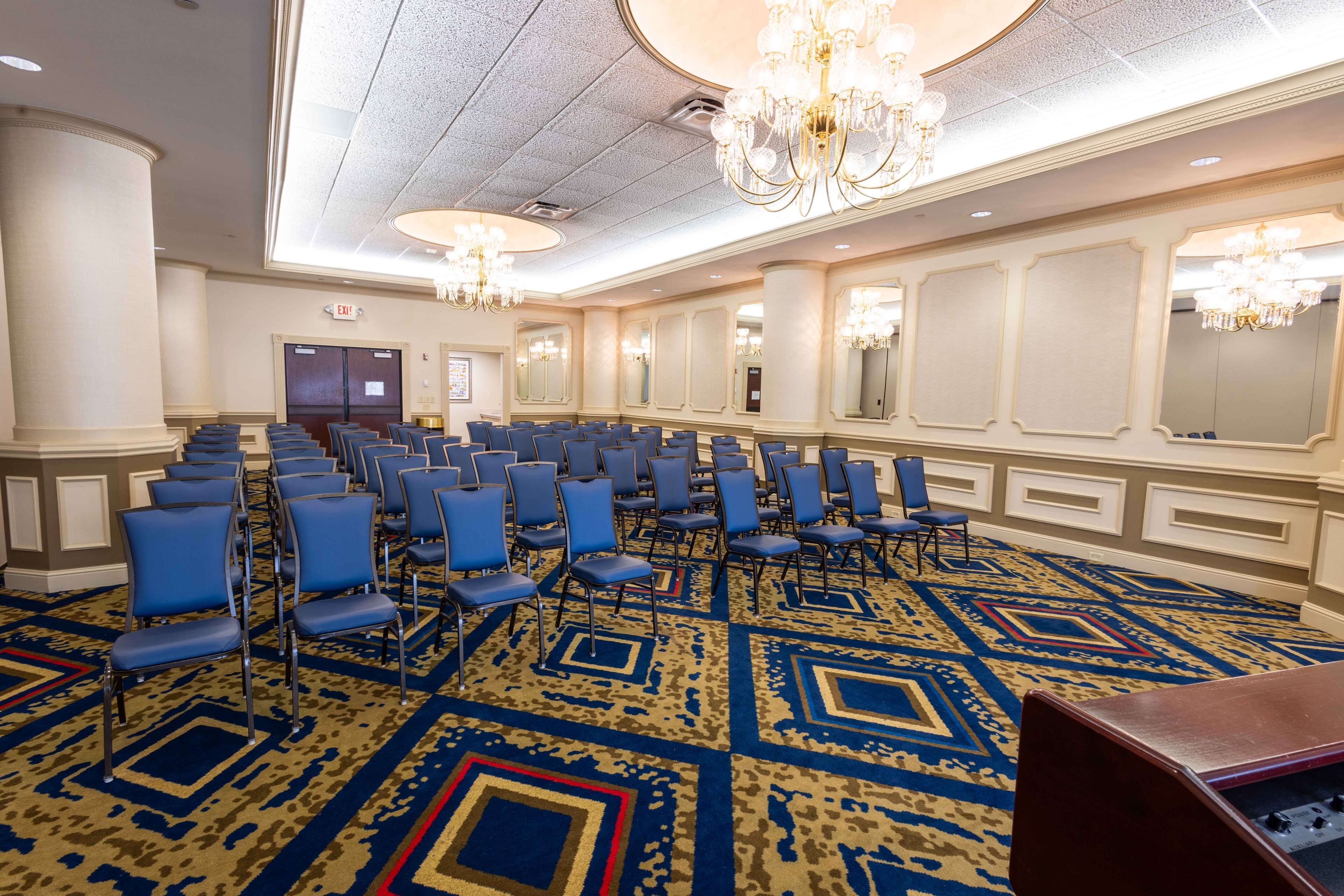 Un amplio salón de conferencias con filas de sillas azules, una alfombra estampada y candelabros.