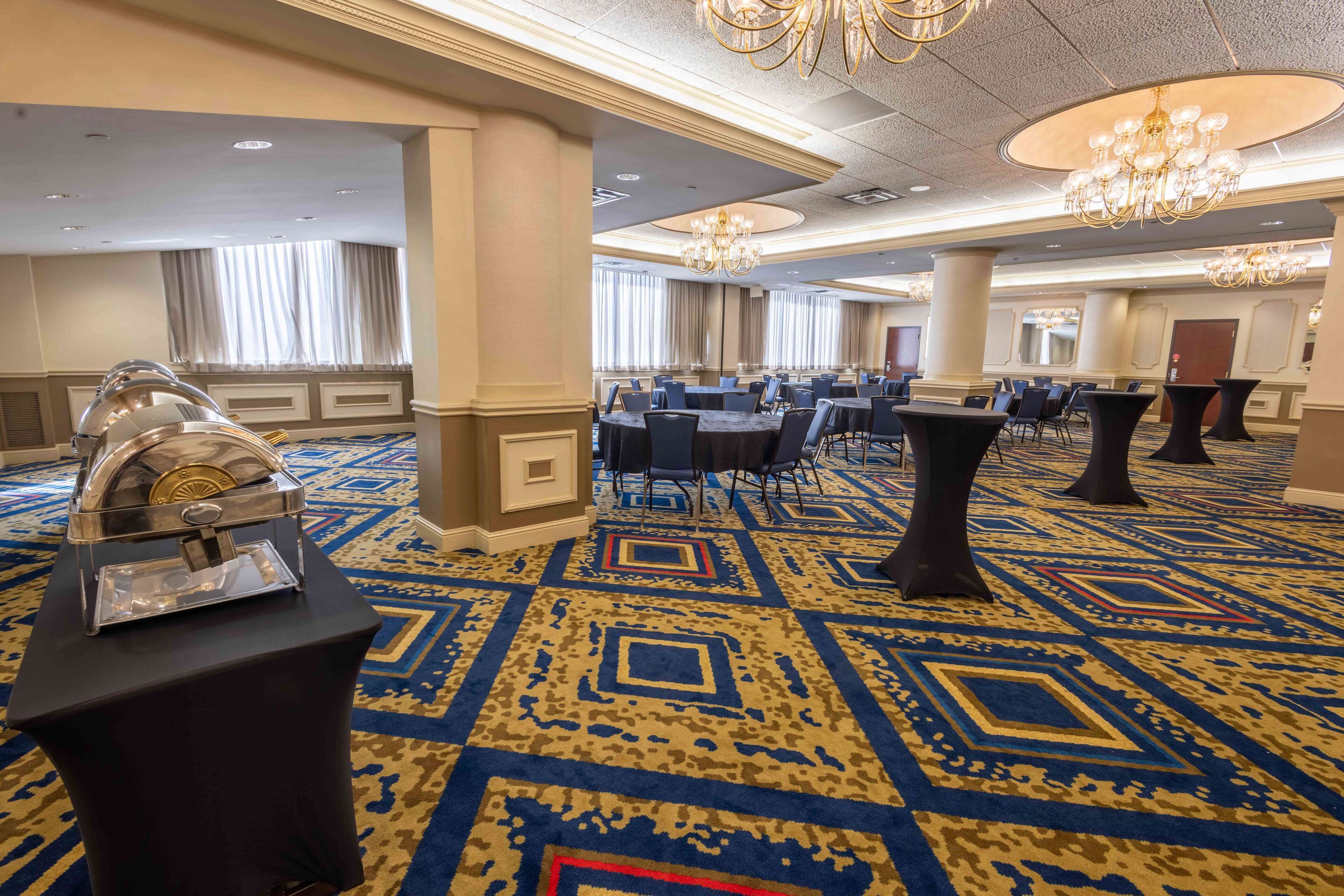 A spacious hotel lobby with a patterned carpet, round tables, and chairs, and a buffet setup.