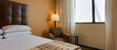 A hotel room with a large bed, a chair, a small table, and a window with curtains.