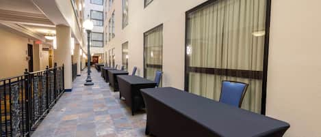 A corridor with a tiled floor, black tables, and chairs, leading to a room with a large window and curtains.