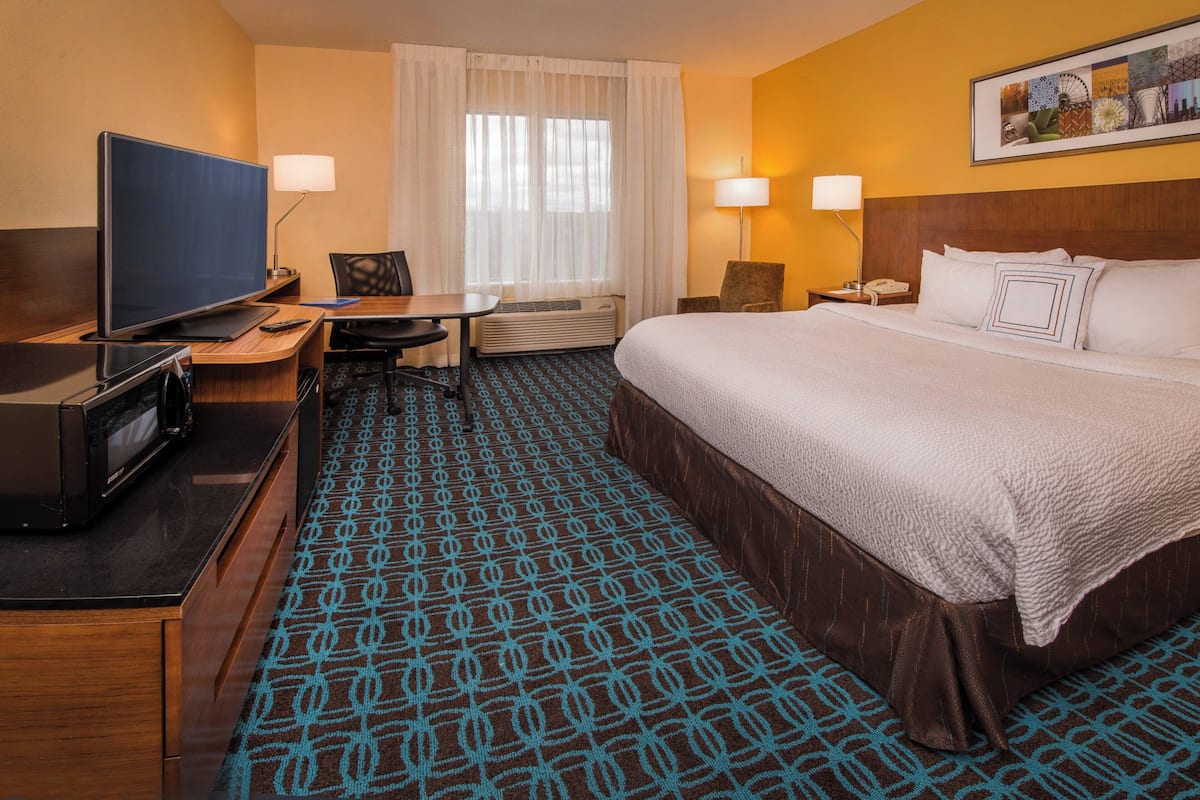 A hotel room with a bed, a desk with a TV, a chair, a table, and a lamp.