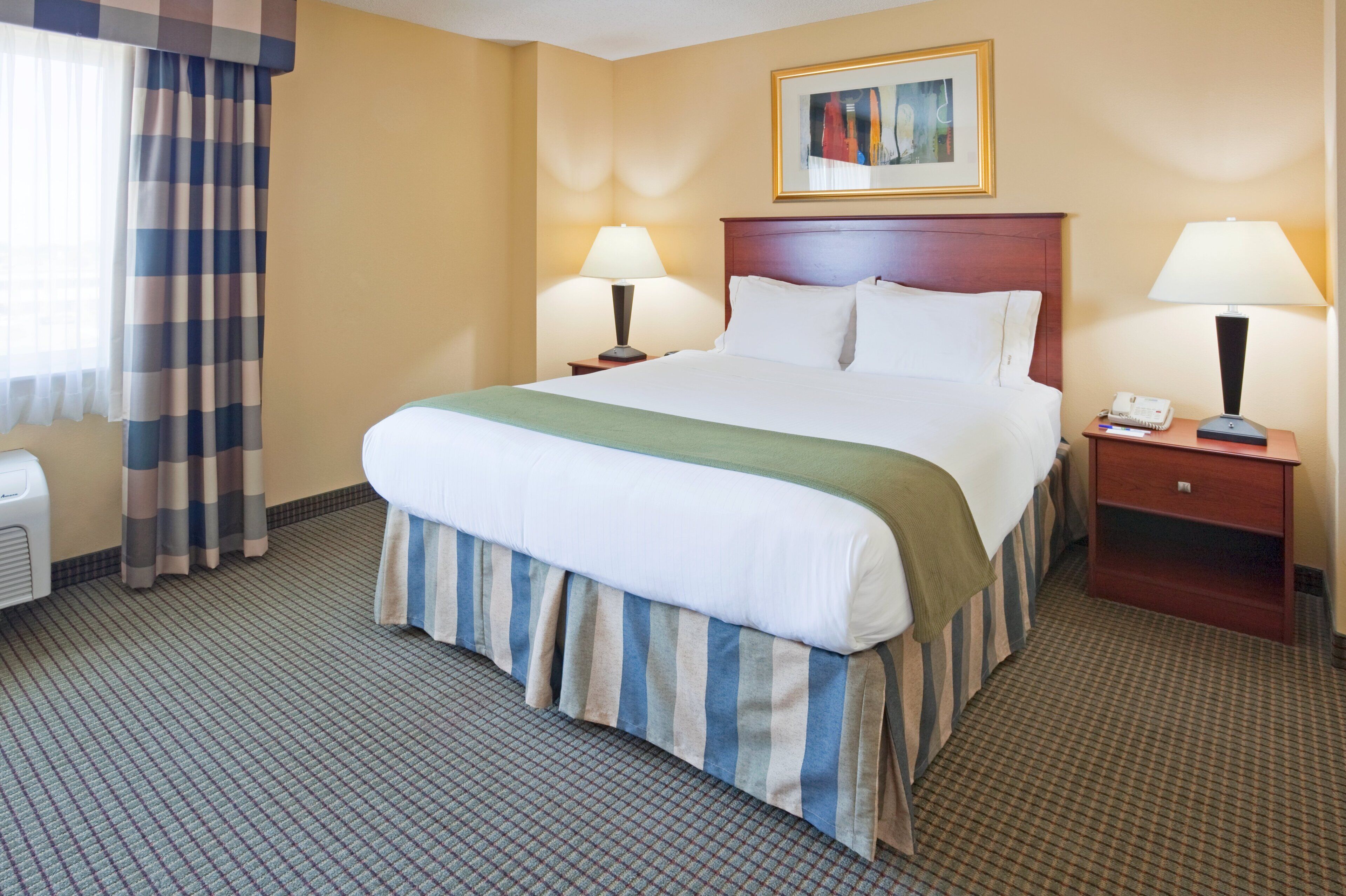 A hotel room with a bed, two bedside tables, a lamp on each table, a painting above the bed, and a window with curtains.