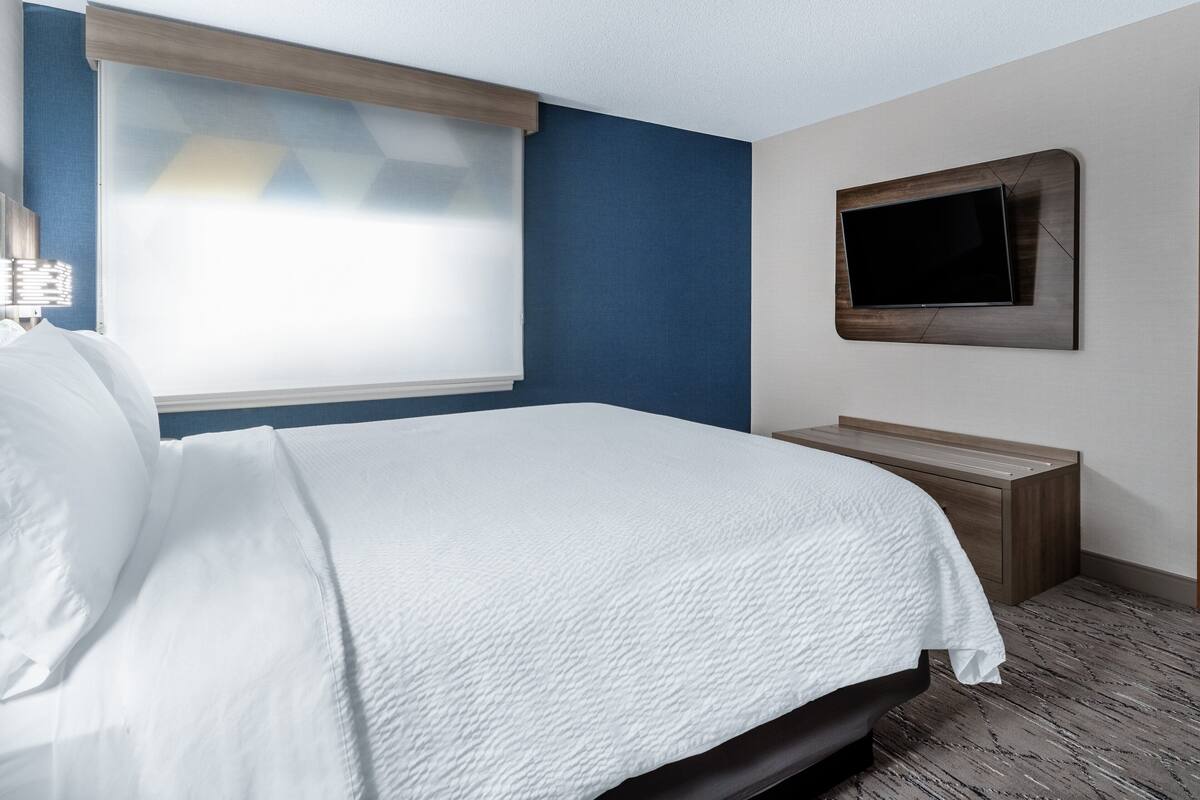 A hotel room with a bed, a television mounted on the wall, and a window with blinds.