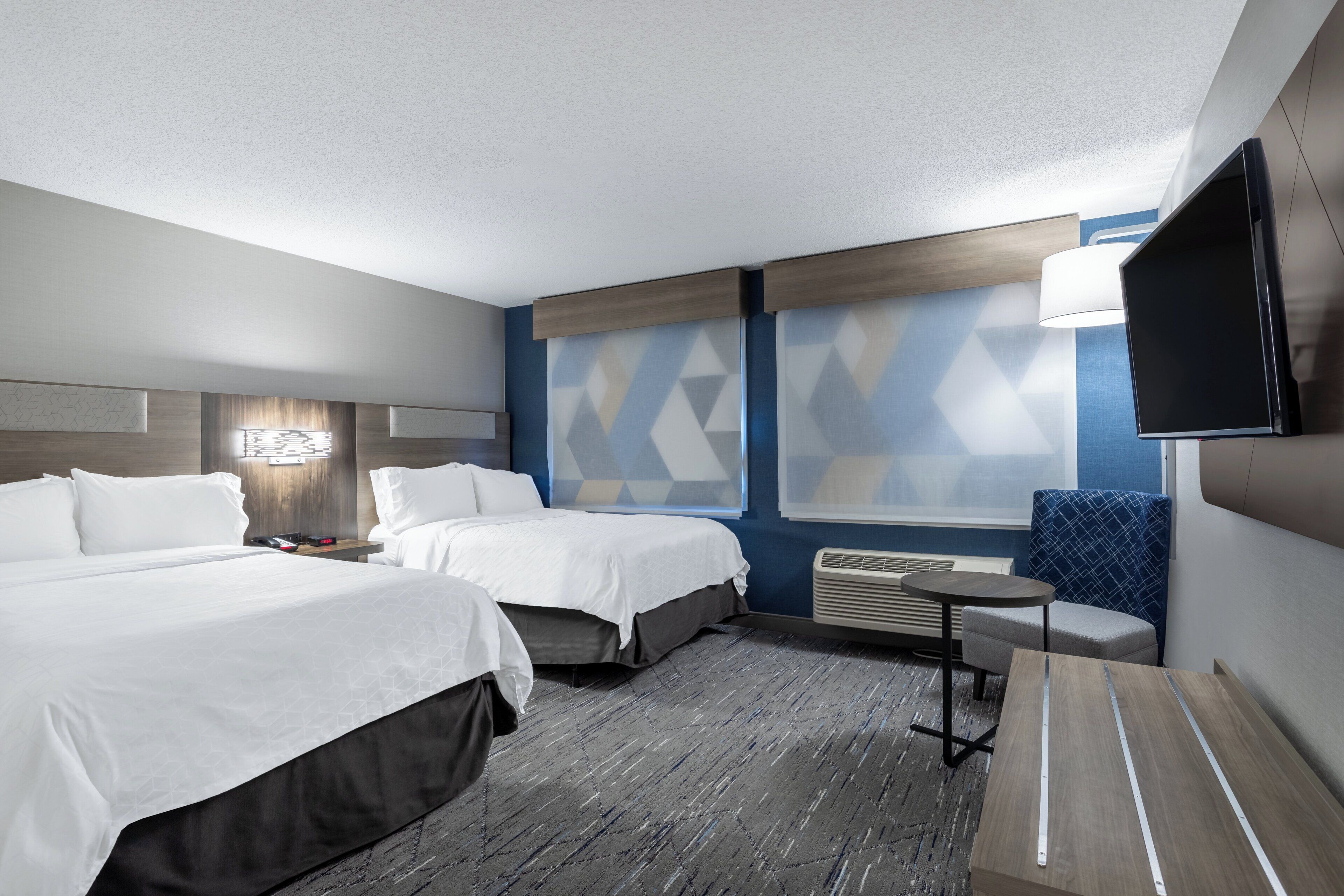 Habitación de hotel con dos camas, un televisor de pantalla plana, una mesita, una silla y una ventana con cortinas de diseño geométrico.
