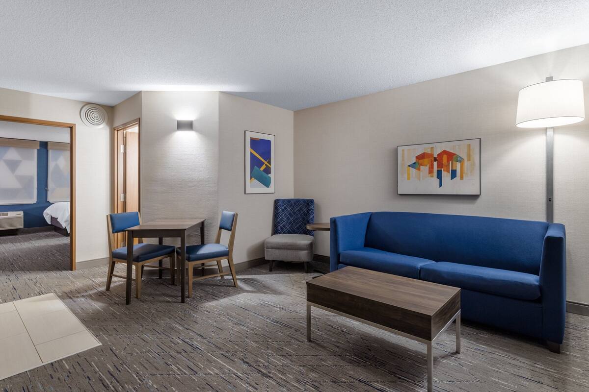 Habitación de hotel con sofá azul, una mesa de centro de madera, una mesa de comedor con sillas y dos lámparas fijadas a la pared.