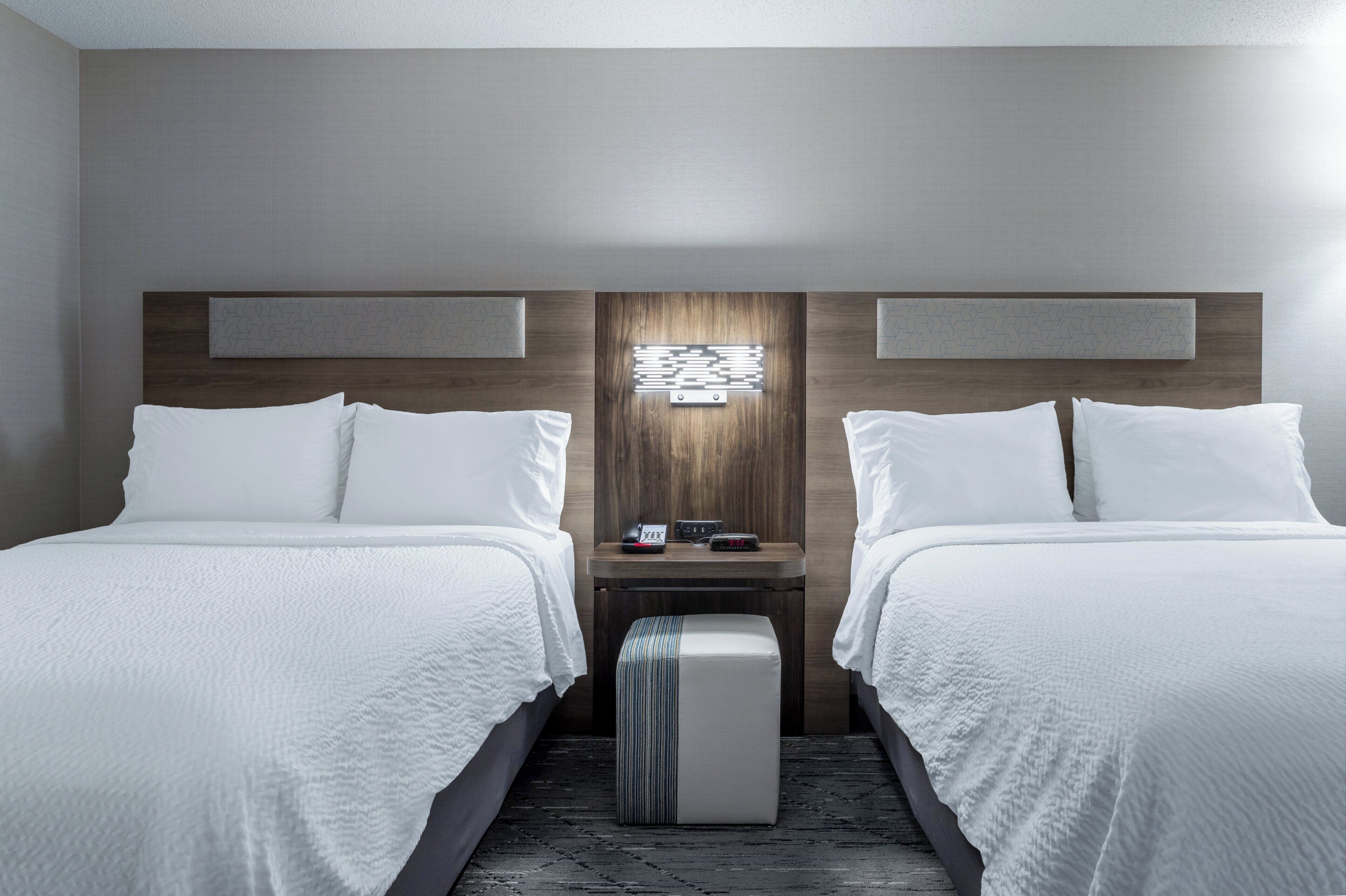 Une chambre d’hôtel avec deux lits, une table de chevet, une lampe fixée au mur et un petit pouf.