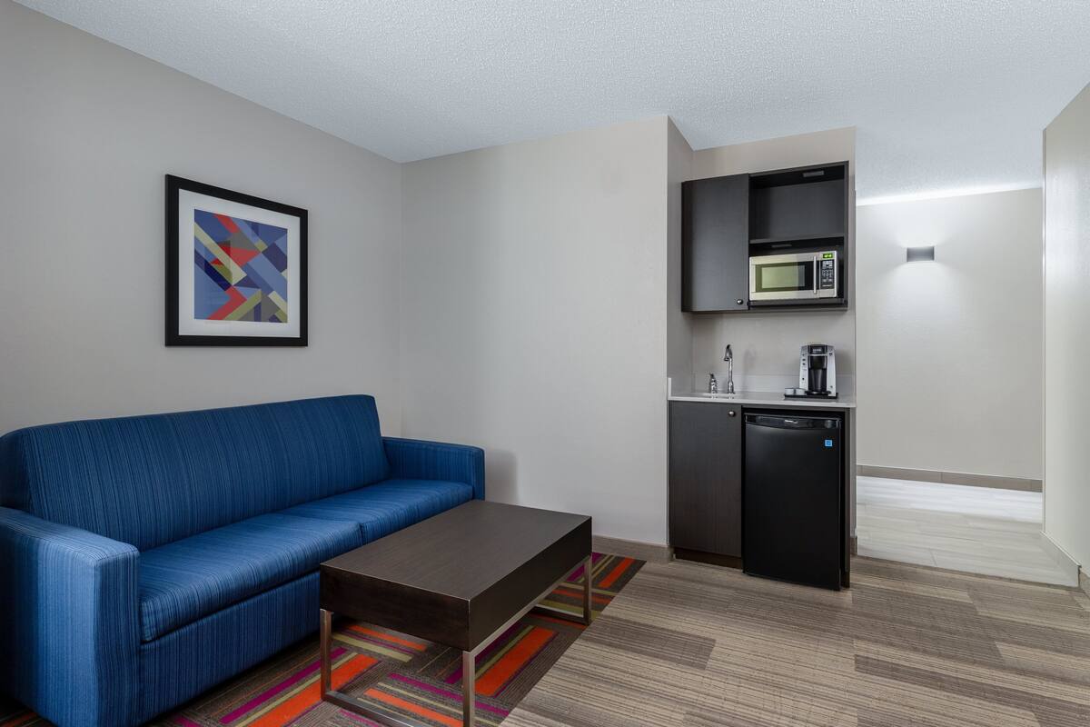 A modern hotel room with a blue sofa, a small coffee table, a colorful rug, a microwave, and a compact refrigerator.