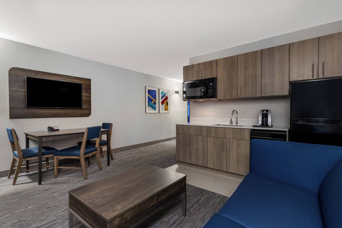 Una sala moderna con sofá, una mesa de centro, zona de comedor con sillas, televisor de pantalla plana en la pared y área de cocina con microondas, refrigerador y pileta.
