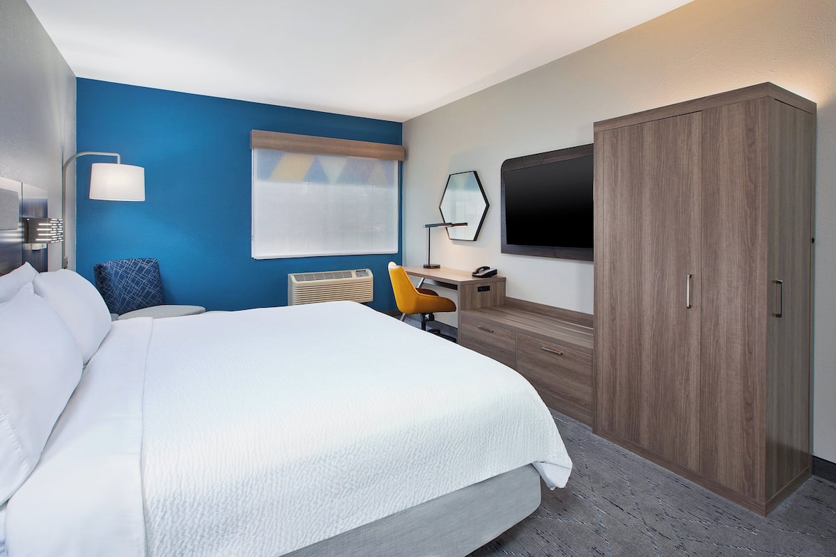 A hotel room with a large bed, a desk with a chair, a wardrobe, a TV, and a window with blinds.